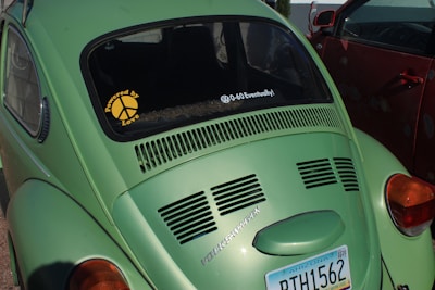 A green Volkswagen Beetle is parked next to a red car. The car displays a sticker on the rear window reading 'Powered by Love' with a peace sign and another message saying '0-60 Eventually!'. An Arizona license plate is visible with the number RTH1562.