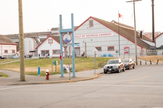 A historic cannery building with red and white coloring stands prominently in the background, featuring signage for the Gulf of Georgia Cannery. Several people are visible near the entrance and along the pathway. Three cars are parked on the street in front, with greenery and walkways surrounding the area. A Canadian flag is flying on a pole, and there is a mix of industrial and rustic architectural elements.