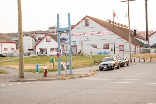A historic cannery building with red and white coloring stands prominently in the background, featuring signage for the Gulf of Georgia Cannery. Several people are visible near the entrance and along the pathway. Three cars are parked on the street in front, with greenery and walkways surrounding the area. A Canadian flag is flying on a pole, and there is a mix of industrial and rustic architectural elements.