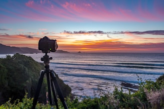 A camera mounted on a tripod is positioned on a grassy cliff overlooking a vast ocean. The sky above is painted with vibrant hues of orange, pink, and purple as the sun sets on the horizon. Gentle waves roll towards the shore against the backdrop of distant islands.