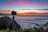 A camera mounted on a tripod is positioned on a grassy cliff overlooking a vast ocean. The sky above is painted with vibrant hues of orange, pink, and purple as the sun sets on the horizon. Gentle waves roll towards the shore against the backdrop of distant islands.