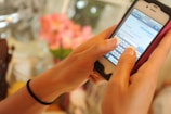 A person is holding a smartphone with a white and black casing, typing a message. The phone screen displays a text conversation in progress. The background is blurred, showing hints of pink flowers.