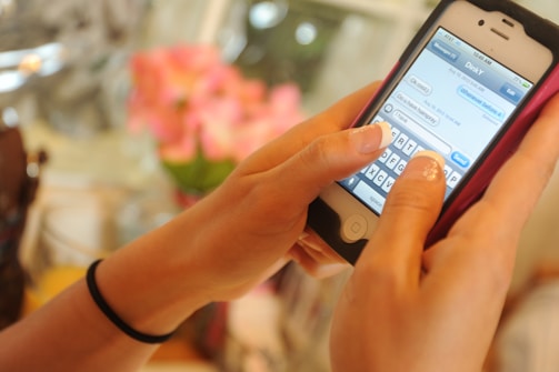 A person is holding a smartphone with a white and black casing, typing a message. The phone screen displays a text conversation in progress. The background is blurred, showing hints of pink flowers.