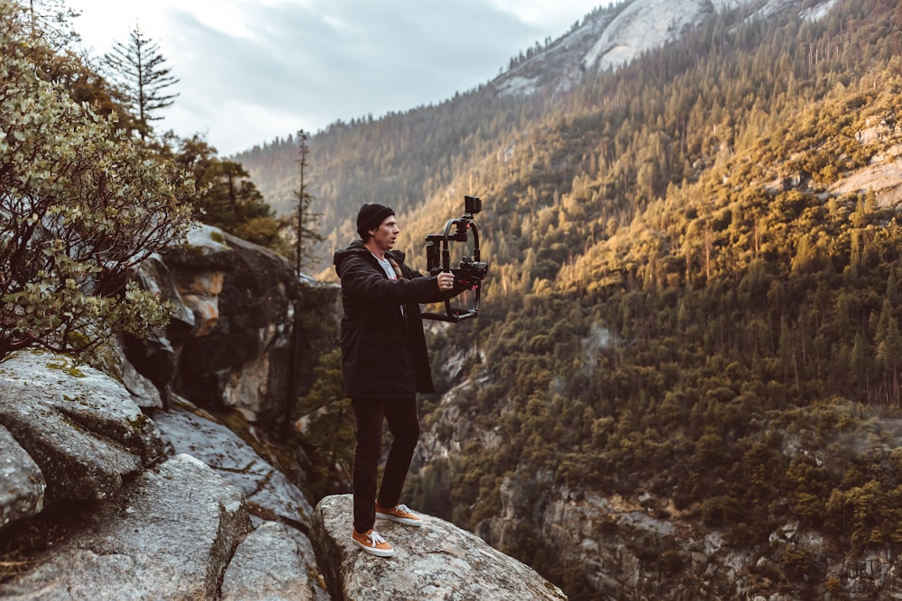 man in black jacket and black pants holding camera standing on rocky mountain during daytime