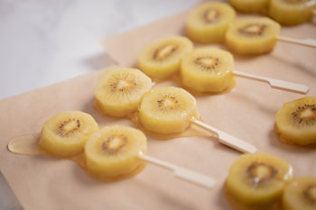 Slices of golden kiwi are arranged on wooden sticks, resembling popsicles, placed on a sheet of parchment paper. The kiwis are glistening, likely coated in a syrup or glaze.