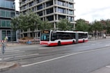 A red and white city bus is traveling on a road in an urban setting. The surroundings include modern buildings with glass facades, trees lining the street, and a barricaded sidewalk area indicating construction or maintenance work. Several bicycles are parked on the sidewalk, and a few pedestrians are visible.