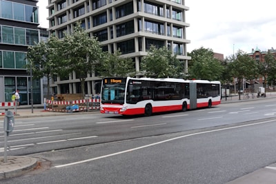 A red and white city bus is traveling on a road in an urban setting. The surroundings include modern buildings with glass facades, trees lining the street, and a barricaded sidewalk area indicating construction or maintenance work. Several bicycles are parked on the sidewalk, and a few pedestrians are visible.