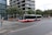 A red and white city bus is traveling on a road in an urban setting. The surroundings include modern buildings with glass facades, trees lining the street, and a barricaded sidewalk area indicating construction or maintenance work. Several bicycles are parked on the sidewalk, and a few pedestrians are visible.