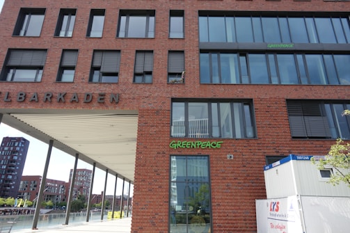 A brick building with large windows features the word 'Greenpeace' in green letters. The building is situated near a waterfront, with other modern buildings visible in the background. There is a white container in the foreground with some text and a tree to the right.