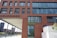 A brick building with large windows features the word 'Greenpeace' in green letters. The building is situated near a waterfront, with other modern buildings visible in the background. There is a white container in the foreground with some text and a tree to the right.