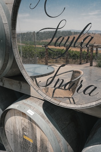 Several wooden barrels are stacked outdoors. One barrel has a glass face with elegant script lettering. The landscape in the background includes rows of grapevines and hills under a clear sky.