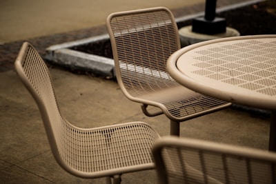 A set of outdoor metal mesh chairs and a round table are arranged on a bare concrete pavement. The design of the furniture is minimalist, with a focus on utility. Surroundings include a planter and a lamp post.