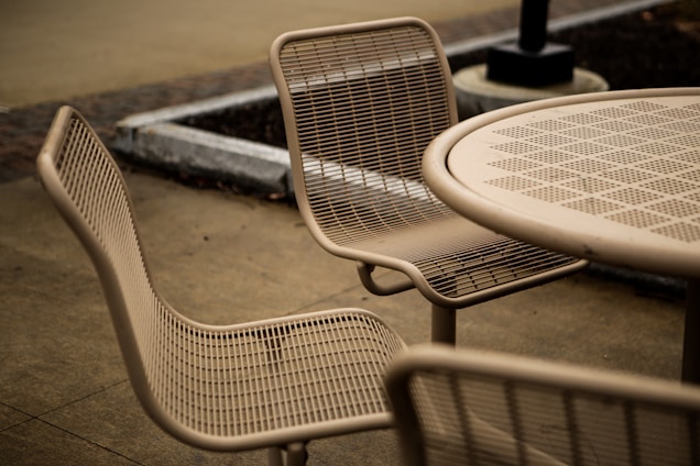 A set of outdoor metal mesh chairs and a round table are arranged on a bare concrete pavement. The design of the furniture is minimalist, with a focus on utility. Surroundings include a planter and a lamp post.