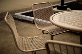 A set of outdoor metal mesh chairs and a round table are arranged on a bare concrete pavement. The design of the furniture is minimalist, with a focus on utility. Surroundings include a planter and a lamp post.