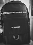 A black backpack with a sleek, modern design. It has a glossy finish and features white text that reads 'PROFESSIONAL' near the top and 'LILIBAIHE' on the pocket flap. The backpack includes a zipper and a metal loop at the bottom of the pocket.