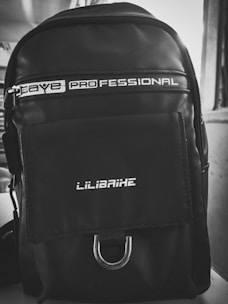 A black backpack with a sleek, modern design. It has a glossy finish and features white text that reads 'PROFESSIONAL' near the top and 'LILIBAIHE' on the pocket flap. The backpack includes a zipper and a metal loop at the bottom of the pocket.