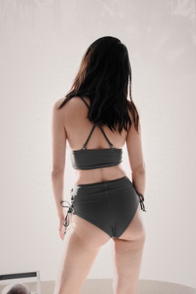 A person with dark hair is wearing a dark green two-piece swimsuit. The swimsuit features a strappy top and high-waisted bottoms with lace-up sides. The person is posing with their back turned to the camera in a minimalist setting.