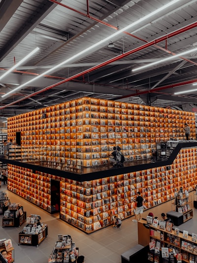 A large, modern bookstore with an eye-catching design features a cube-shaped structure filled with shelves of books. The walls are illuminated with warm lighting, creating a cozy and inviting atmosphere. People are browsing through the books on multiple levels, and the spacious interior is filled with various stands displaying more reading material.