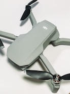 A compact, modern drone with a sleek grey body and four black propellers. The drone is positioned on a light surface, showcasing its aerodynamic design.