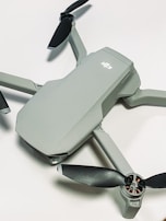 A compact, modern drone with a sleek grey body and four black propellers. The drone is positioned on a light surface, showcasing its aerodynamic design.