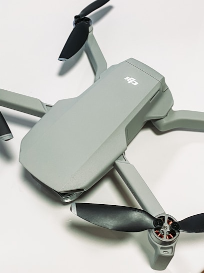 A compact, modern drone with a sleek grey body and four black propellers. The drone is positioned on a light surface, showcasing its aerodynamic design.