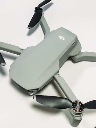 A compact, modern drone with a sleek grey body and four black propellers. The drone is positioned on a light surface, showcasing its aerodynamic design.
