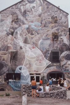 A large mural covers the side of a multi-story building, depicting scenes of pottery and ceramics with detailed imagery of various jars, vases, and people engaged in artistic activities. People are gathered in front, sitting or standing near stone benches, observing the artwork.