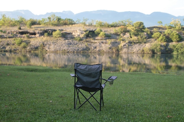 A solitary folding camping chair is placed on a grassy lawn, facing a calm body of water with reflections of the surrounding terrain. Beyond the water, a rocky embankment with scattered green vegetation can be seen, with distant mountains under a clear sky in the background.