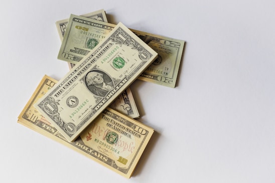 Several US dollar bills of various denominations are spread over a plain surface. The bills include one, ten, and twenty dollar notes, stacked irregularly on a neutral backdrop.