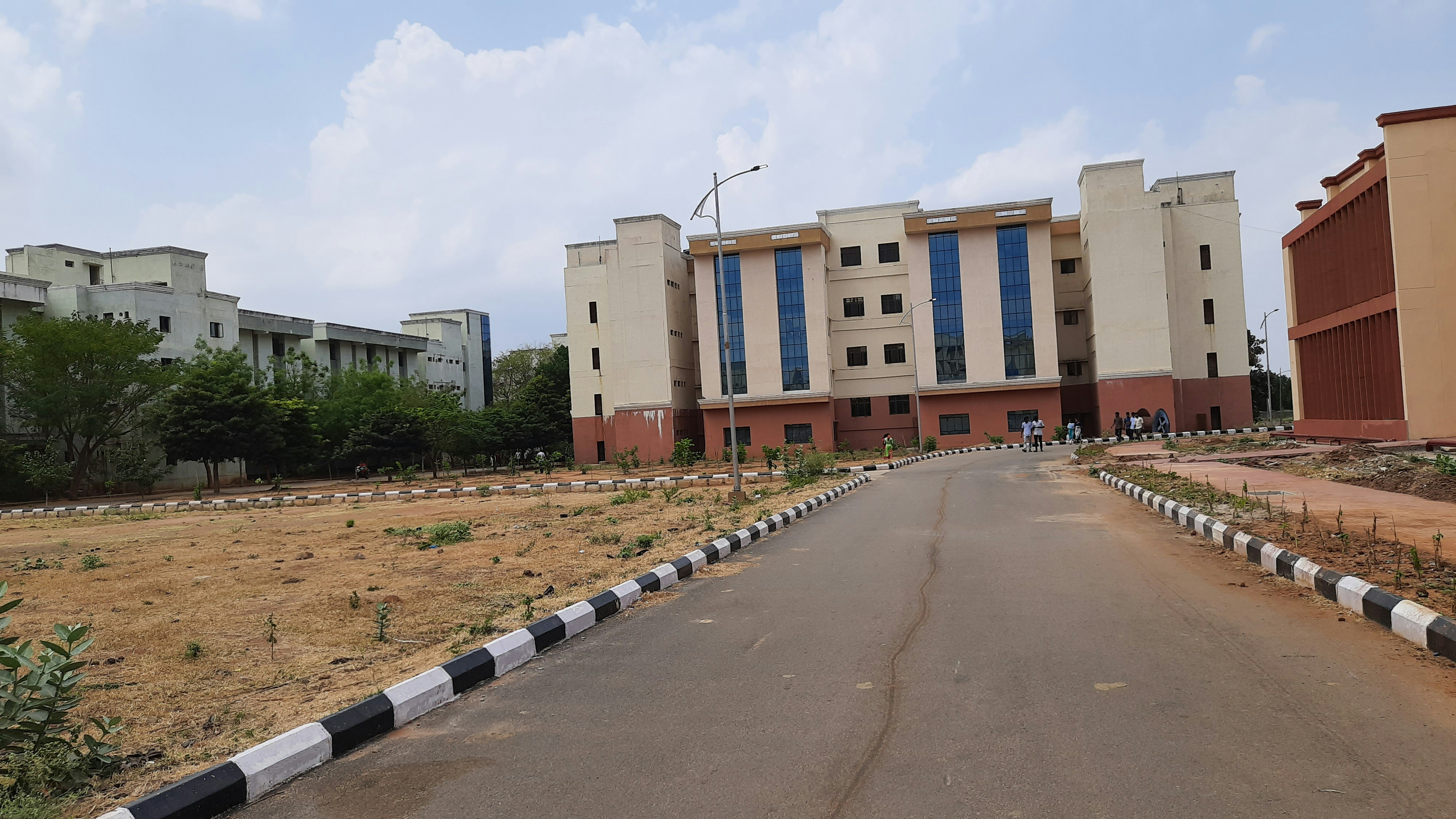 A multi-story educational or institutional building with large blue-tinted windows, surrounded by a paved road and some vegetation. The foreground features a landscaped area with young plants and a black-and-white painted curb. Several people are gathered near the entrance of the building.