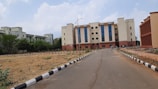 A multi-story educational or institutional building with large blue-tinted windows, surrounded by a paved road and some vegetation. The foreground features a landscaped area with young plants and a black-and-white painted curb. Several people are gathered near the entrance of the building.