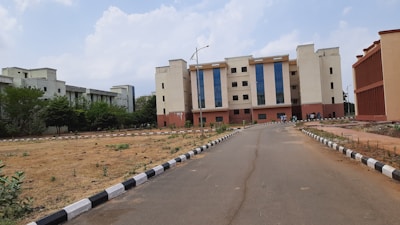 A multi-story educational or institutional building with large blue-tinted windows, surrounded by a paved road and some vegetation. The foreground features a landscaped area with young plants and a black-and-white painted curb. Several people are gathered near the entrance of the building.