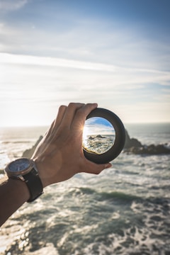 A hand wearing a watch is holding a lens or glass filter against a backdrop of a seascape. The lens magnifies a portion of the ocean and rocky shore, creating a focal point that contrasts with the broader view of the sea and sky. The lighting suggests a time near sunset or sunrise, with warm tones and a bright sun casting reflections on the water.