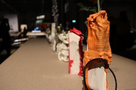 A row of objects placed on a tabletop, each wrapped in a unique material such as an orange rubber material with some text and diagrams on it. The setting appears to be indoors with a dimly lit environment and blurred figures in the background.
