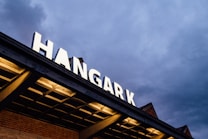 An illuminated sign spelling 'HANGAR' on a building, set against a cloudy evening sky. The structure displays a combination of modern and industrial design elements, with visible grid patterns and warm lighting from beneath the overhang.