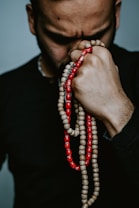 A person with a solemn expression holds a string of prayer beads tightly against their forehead. The beads are a combination of beige and vibrant red colors, and the person's hand is prominently featured.