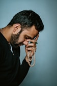 A person with a contemplative expression holds a string of prayer beads. The individual has a hand placed on their forehead, suggesting deep thought or introspection. The background is plain and softly colored.