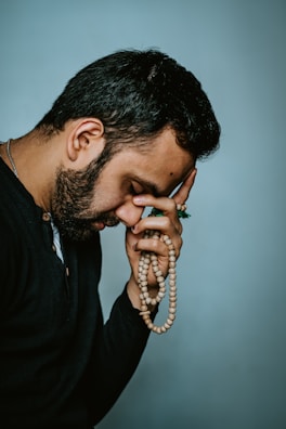 A person with a contemplative expression holds a string of prayer beads. The individual has a hand placed on their forehead, suggesting deep thought or introspection. The background is plain and softly colored.
