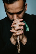 A person with a beard is holding prayer beads in their hands, which are clasped together. The individual appears to be deep in thought or prayer. The image is closely cropped, focusing on the upper body and hands.