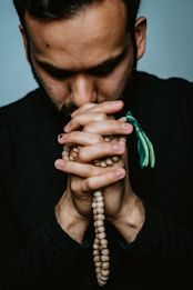 A person with a beard is holding prayer beads in their hands, which are clasped together. The individual appears to be deep in thought or prayer. The image is closely cropped, focusing on the upper body and hands.