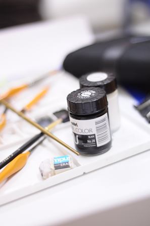 Various art supplies are arranged on a white surface, including black poster color jars, paintbrushes with orange handles, and an eraser. The background is softly blurred, indicating a creative workspace.