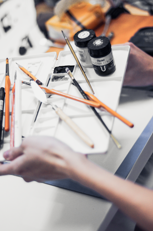 A close-up of various art supplies, including paintbrushes, a mixing palette, and two jars of paints, being held by a pair of hands. The supplies are arranged on a white tray, with some orange-handled brushes and metal palette knives visible.
