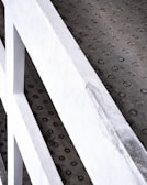 A close-up view of a textured concrete surface with evenly spaced circular indentations. A partial view of white painted metal railings runs diagonally across the image.