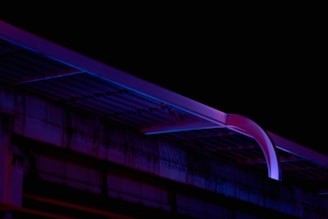 A dimly lit scene featuring part of a building structure illuminated by neon lights in shades of blue and purple. The lighting casts a futuristic and mysterious vibe over the otherwise dark surroundings, while the architecture showcases clean, angular lines.