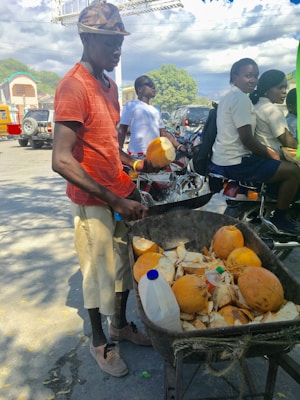 A man wearing a red shirt and cap is standing beside a wheelbarrow filled with coconuts and coconut husks. In the background, there are people sitting on motorcycles and some parked vehicles. The scene takes place outdoors on a sunny day, with a few trees and a building visible in the background.