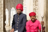 Two individuals wearing traditional attire, including vibrant red turbans, are seated in front of an intricately designed architectural backdrop. One person is wearing a dark colored jacket with brass buttons, while the other is in a bright pink shirt holding an ornate staff. The background features detailed carvings and decorative paneling, suggesting a historical or cultural setting.