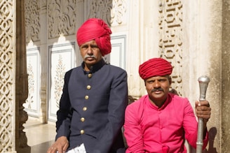 Two individuals wearing traditional attire, including vibrant red turbans, are seated in front of an intricately designed architectural backdrop. One person is wearing a dark colored jacket with brass buttons, while the other is in a bright pink shirt holding an ornate staff. The background features detailed carvings and decorative paneling, suggesting a historical or cultural setting.