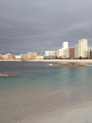 A coastline with clear turquoise waters gently touching a sandy beach in the foreground. In the background, a cityscape of modern high-rise buildings lines the shore under a cloudy sky.