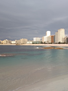 A coastline with clear turquoise waters gently touching a sandy beach in the foreground. In the background, a cityscape of modern high-rise buildings lines the shore under a cloudy sky.