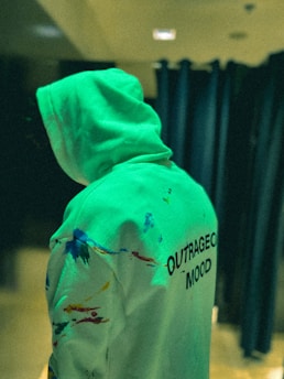 A person wearing a hoodie with colorful paint splatters and the words 'OUTRAGEOUS MOOD' printed on the back, standing in a dimly lit environment with dark curtains in the background.